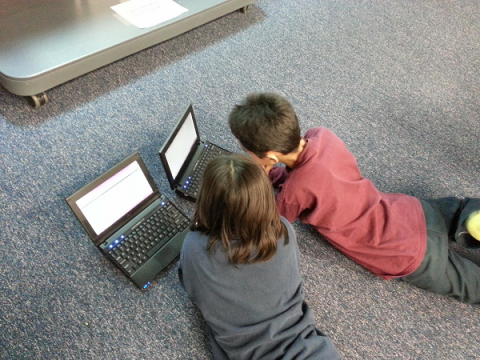Two primary school aged children, lying on the floor on their bellies, using a laptop each